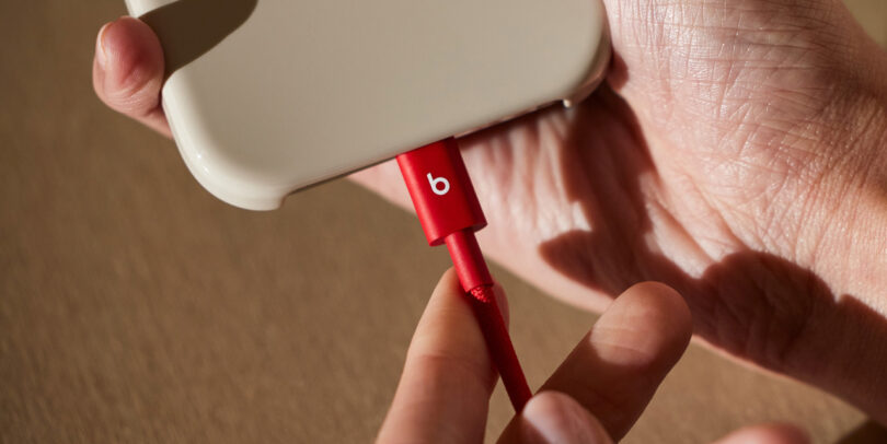 A person plugs a red Beats charging cable into a beige smartphone case, with hands visible holding both the device and the distinctive Beats cable.