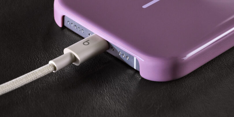 A braided charging cable plugged into a smartphone with a purple case, resting on a black leather surface next to Beats headphones.