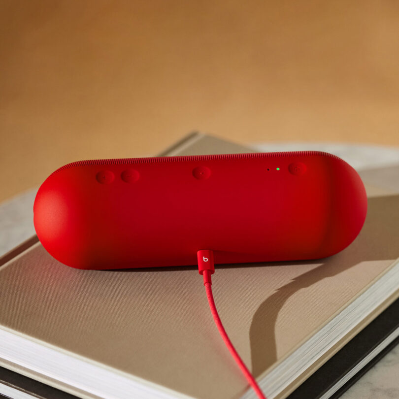A red Beats portable wireless speaker is charging on top of two closed books, with a red charging cable plugged in.