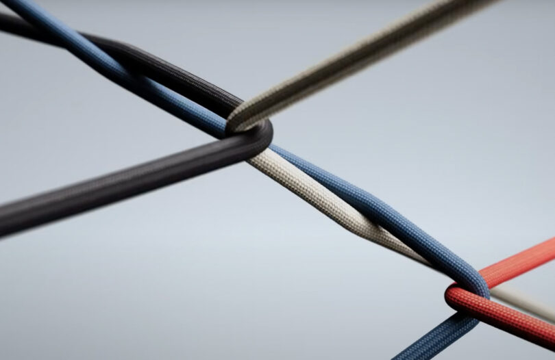 Four colored cords—black, blue, beige, and red—are intertwined in a geometric pattern against a plain, light background, evoking the rhythmic movement of beats.