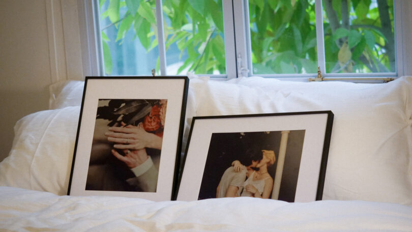 Two framed photographs rest against white pillows on a bed, with a window and green leaves visible in the background.