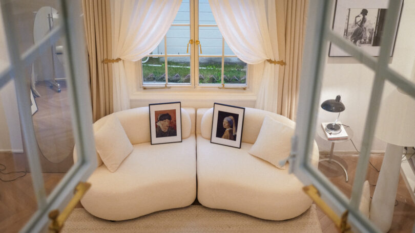 A cozy living room with a white curved sofa, two framed portraits on the sofa, a window with sheer curtains, and a side table with a lamp and framed photo.