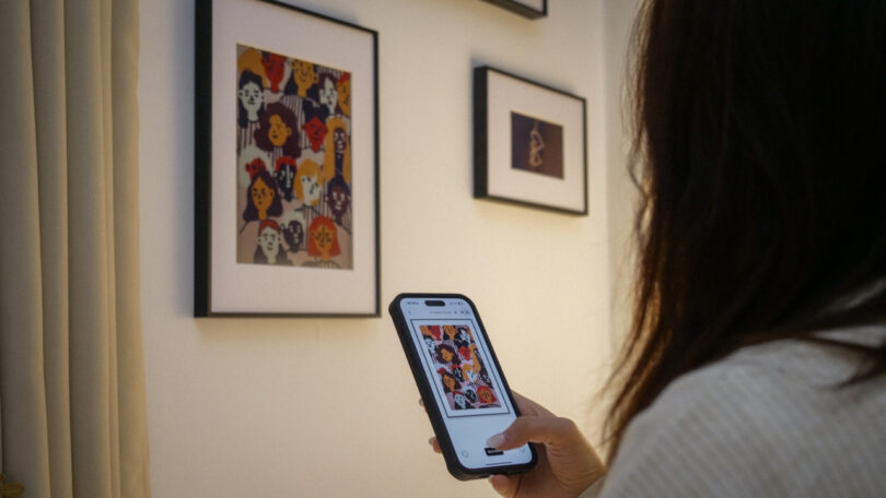 A person holds a smartphone and takes a photo of framed artwork on a wall, focusing on a colorful portrait piece.