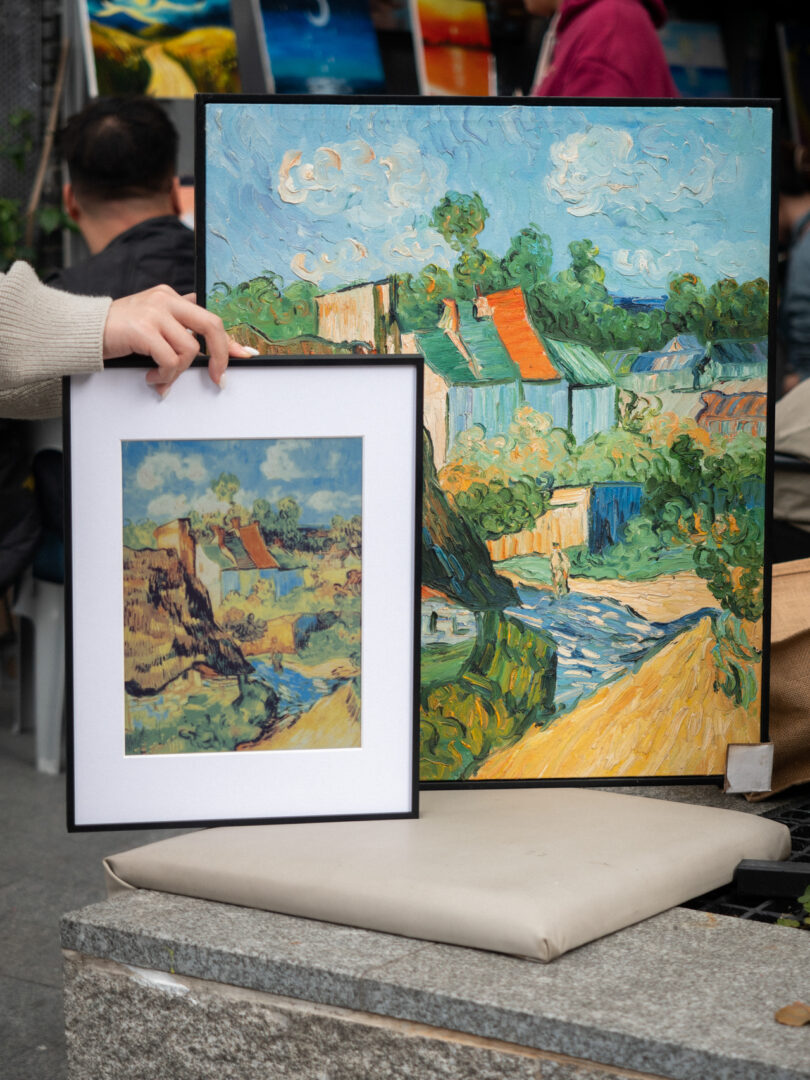 Person holding a framed print of a painting next to a larger, similar painting; both depict a colorful landscape in an impressionist style.