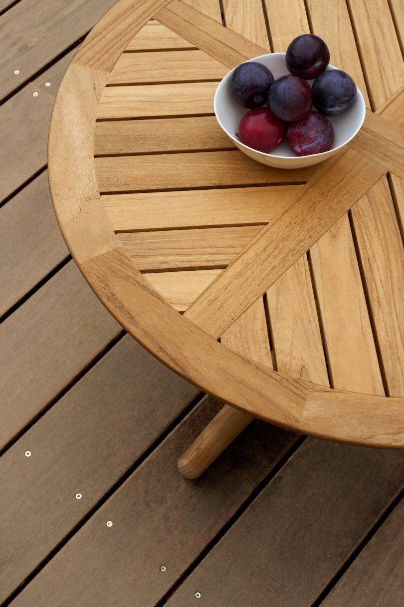 A round wooden table with a white bowl of plums and red fruits on a wooden deck