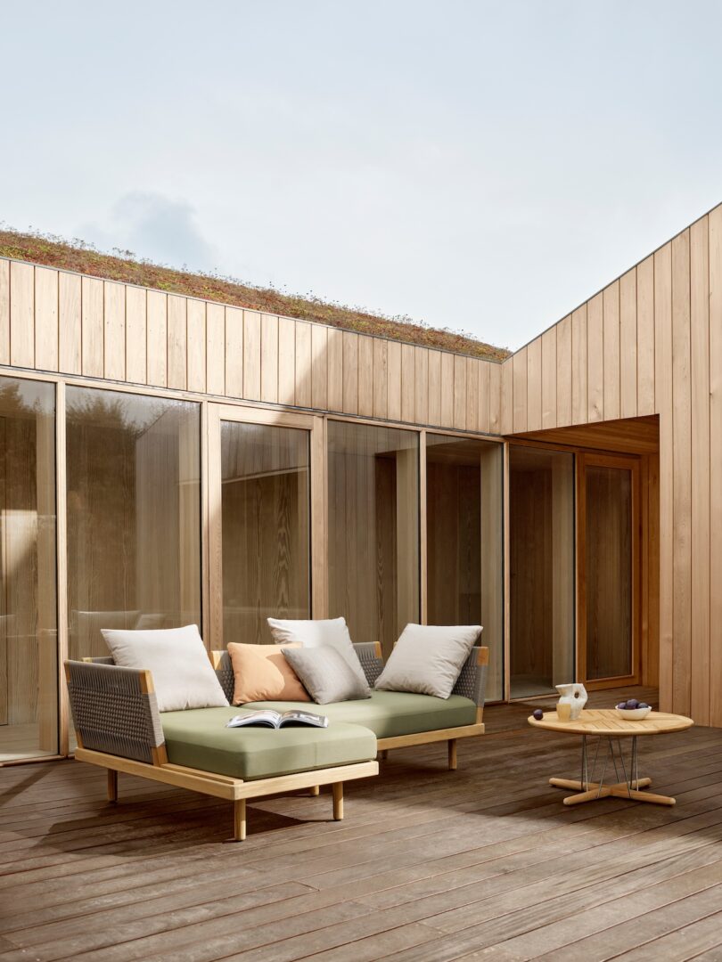 Modern outdoor patio setting with a wooden sofa, green and white cushions, an open book, a small round table, and a mug. The area is surrounded by wooden panel walls and large glass doors