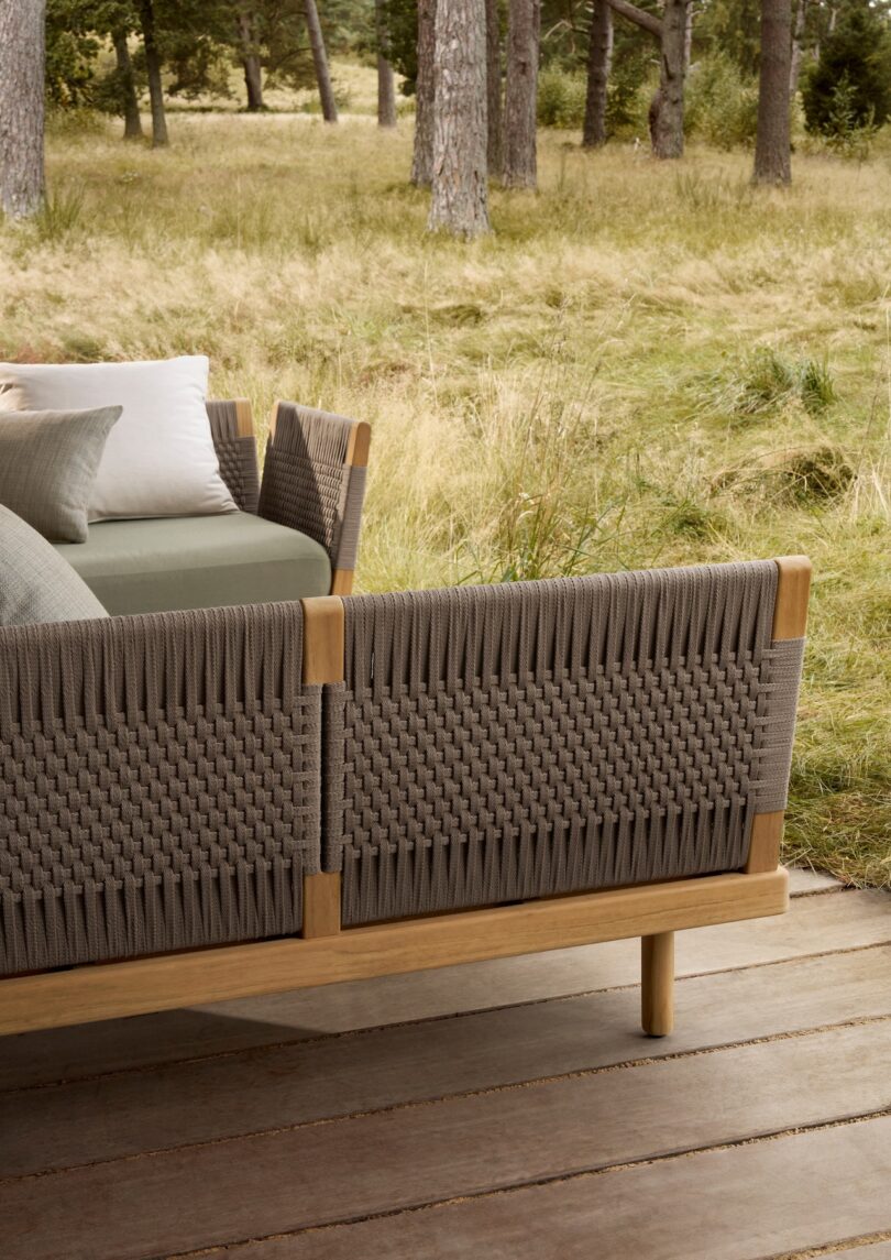 Outdoor wicker sofa with woven backrest and wooden frame on a wooden deck, surrounded by grass and trees in the background