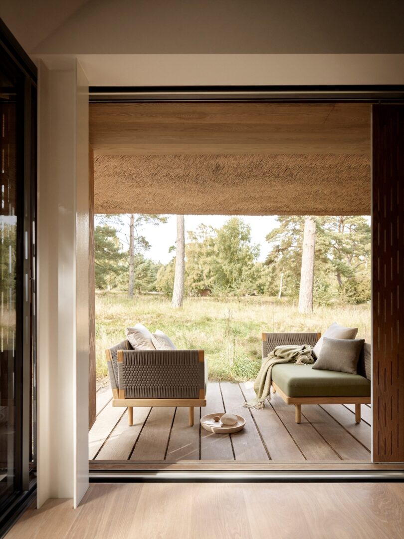 View of a wooden patio with modern outdoor furniture, overlooking a grassy field and trees in the distance. A plate and blanket are on the seating