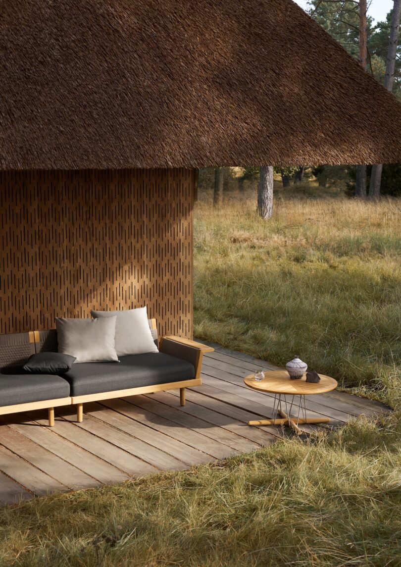 Outdoor wooden deck with a sofa and cushions, small round table, set beneath a thatched roof structure surrounded by grass and trees