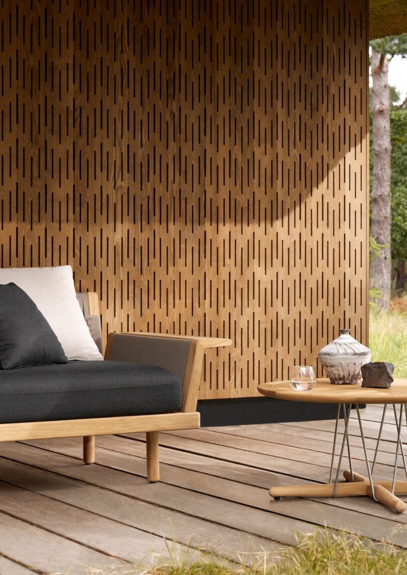 Outdoor seating area with a wooden sofa and cushions, a side table holding a teapot and bowl, situated on a deck with a wooden slat wall in the background. Grass surrounds the deck