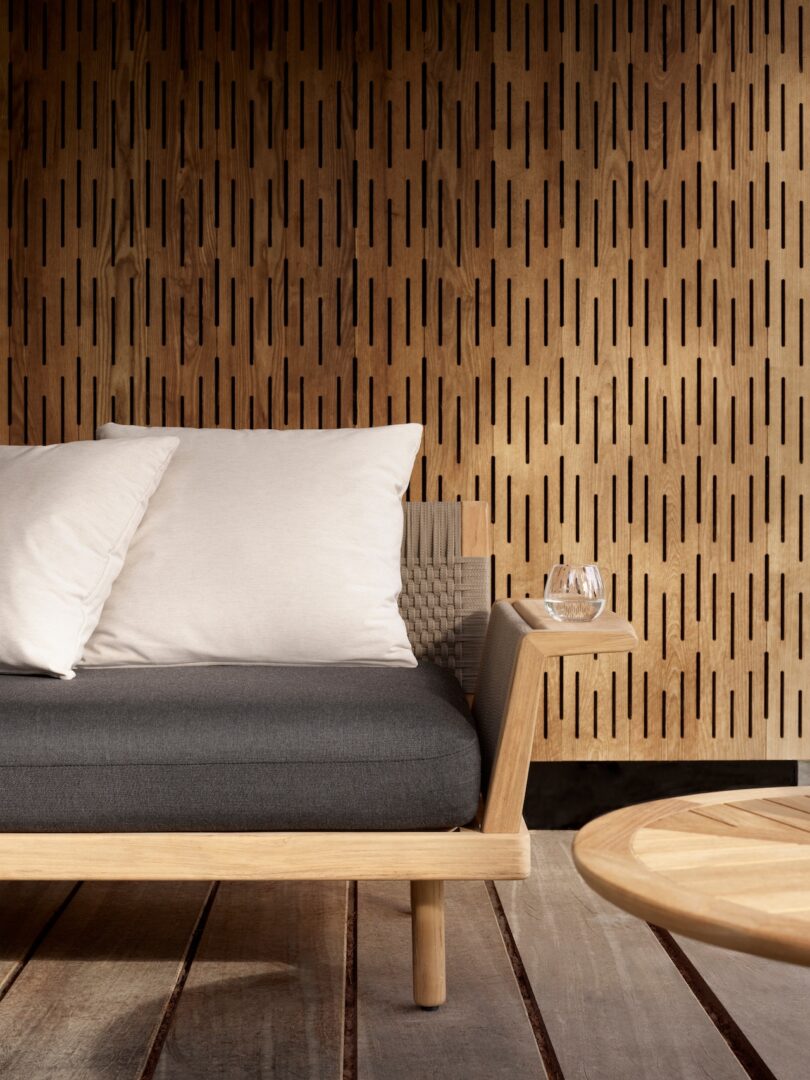 Modern outdoor seating area with a wooden sofa, white cushions, and a round table. A glass of water sits on the sofa's arm. Vertical patterned wooden wall in the background