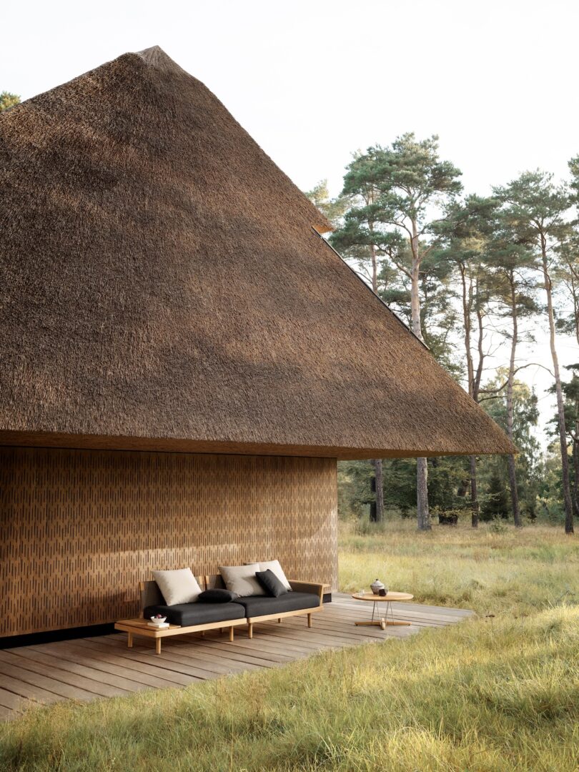 A thatched-roof house with a wooden deck featuring outdoor seating. It is situated in a grassy area with tall trees in the background