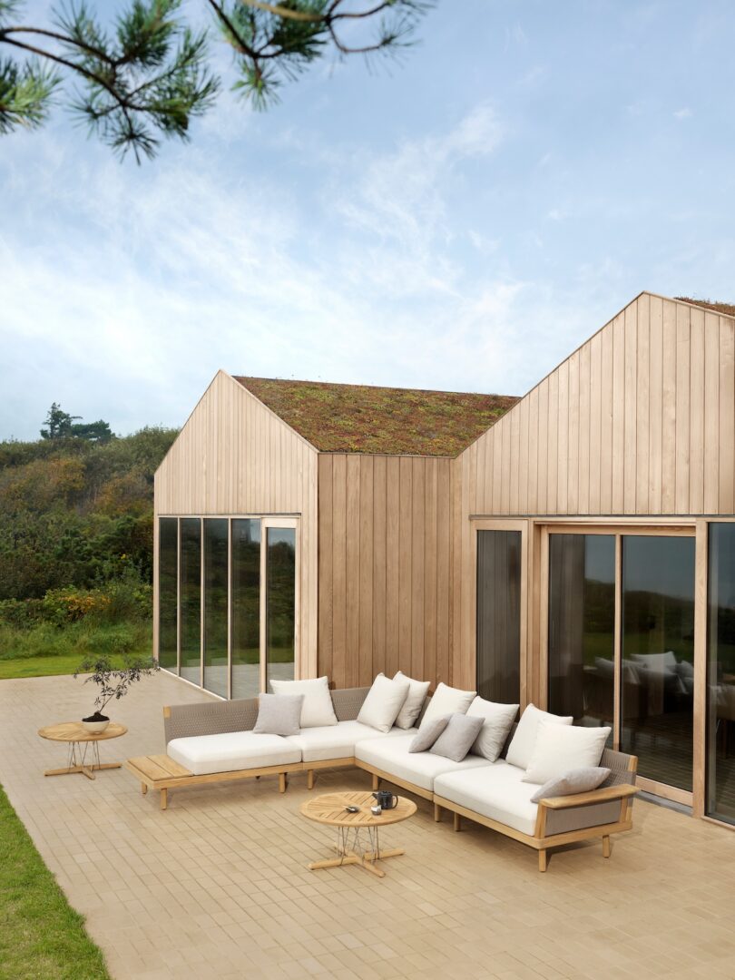 Wooden house with a grass roof, large glass windows, and an outdoor patio featuring white cushioned seating and round wooden tables against a clear sky backdrop