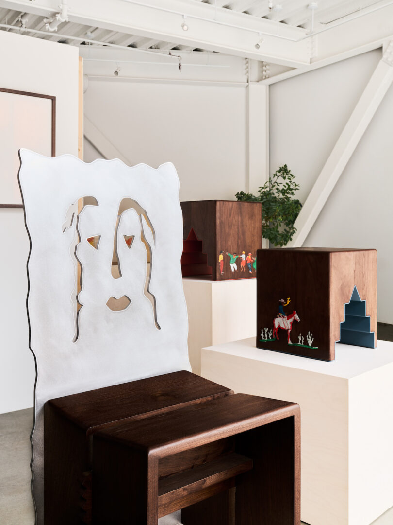 A modern gallery space displays sculptural wooden furniture, including a white chair with a face cutout and wooden cubes with colorful painted figures and shapes