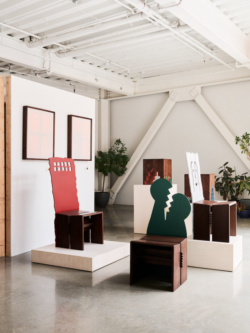 An art gallery space with modern, geometric chairs and stools on display, abstract framed artworks on the wall, and potted plants in the background