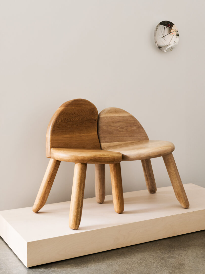Two small wooden chairs with rounded backs and three legs each are joined together, positioned on a light wooden platform with a round wall clock above them
