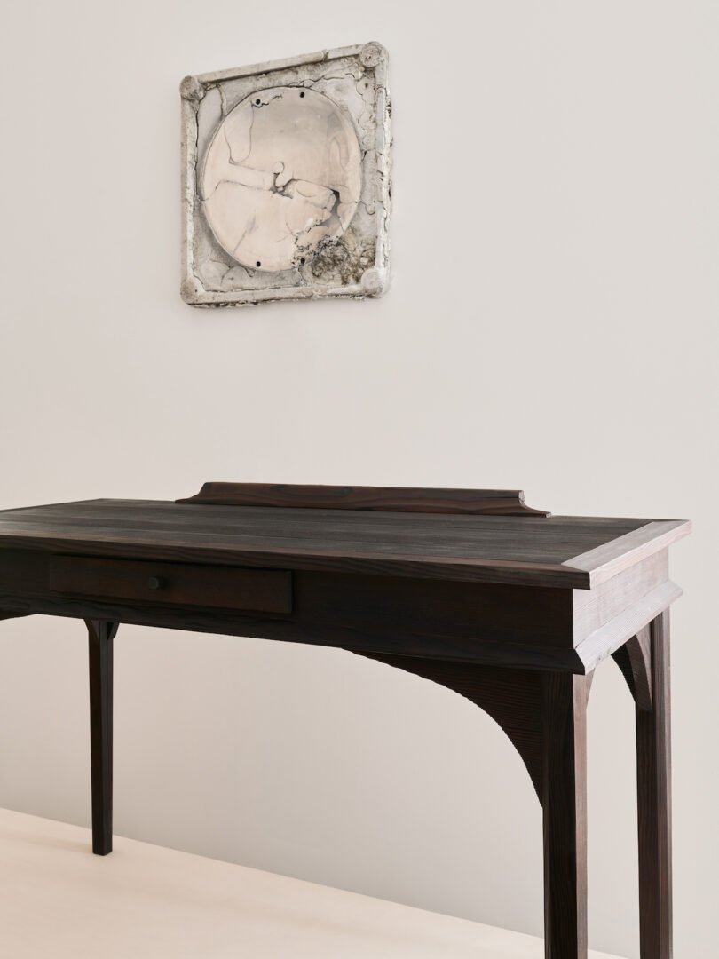 A dark wooden table with a single drawer stands against a light-colored wall, beneath a square abstract artwork in neutral tones