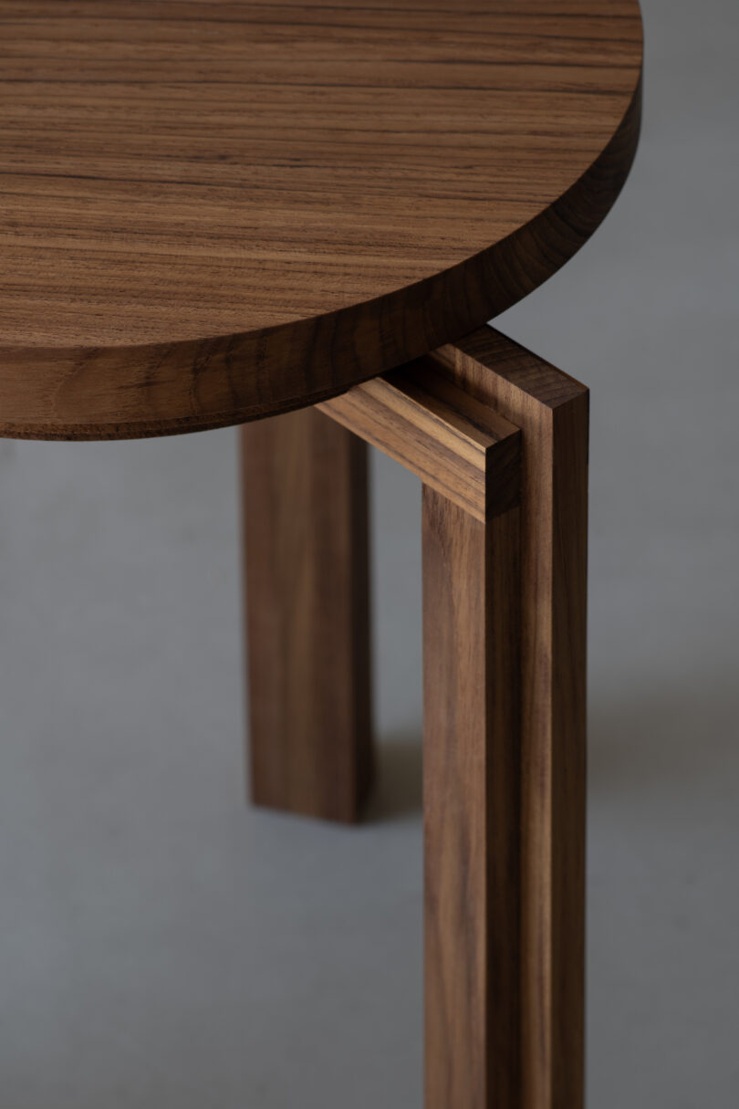 Close-up of a wooden round tabletop with legs. The wood has a natural, dark grain pattern. The focus is on the corner where the leg meets the table