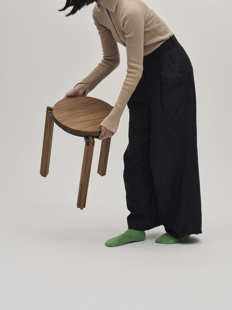 A person in a beige sweater and black pants holds a small wooden stool. They are wearing green socks