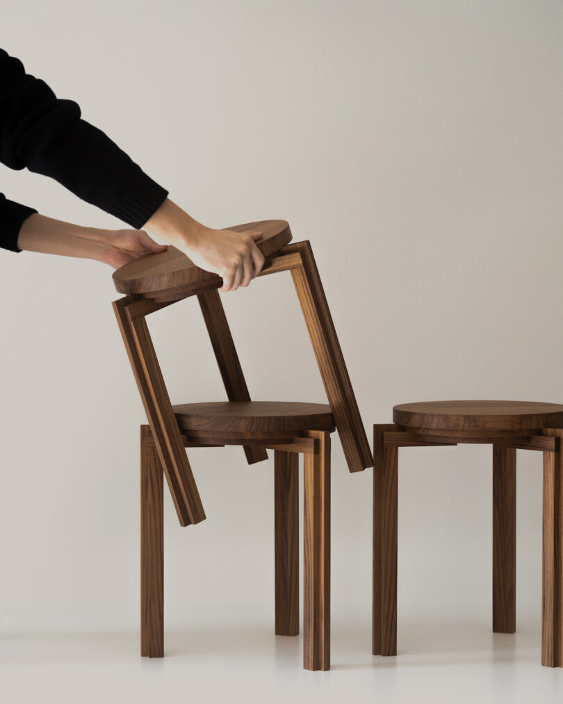 Person stacking wooden stools with round seats and square legs on a white surface