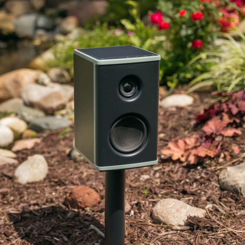A black outdoor speaker mounted on a post surrounded by rocks, mulch, and foliage.