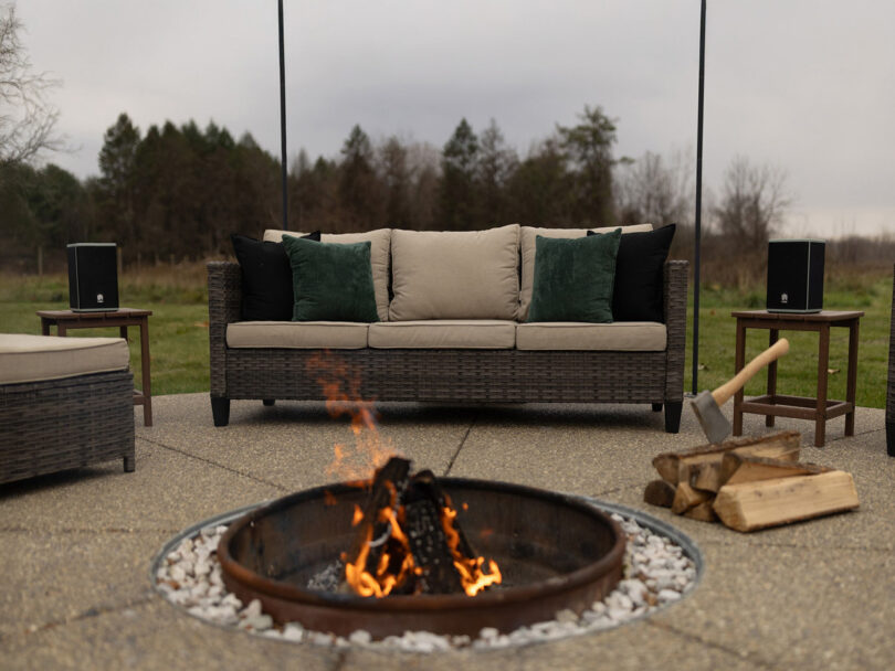 Outdoor seating area with a sofa, cushions, two speakers, and a fire pit. An axe is placed on a log near the fire. Trees and overcast sky in the background.