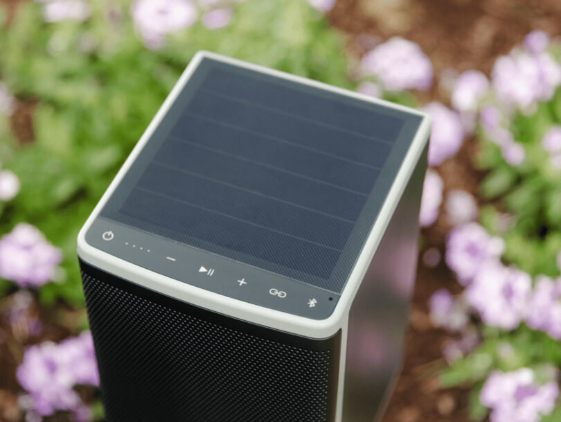Rectangular portable speaker with solar panel top, control buttons, and Bluetooth symbol on the side. Pink flowers and greenery in the blurred background.