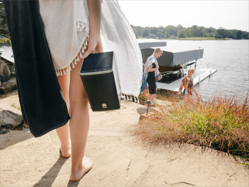 Person holding a speaker while descending steps toward a dock with people by a lake.