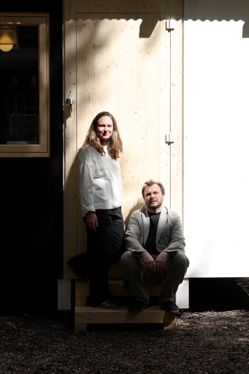 A woman stands and a man sits on wooden steps outside a building, both looking at the camera in natural daylight