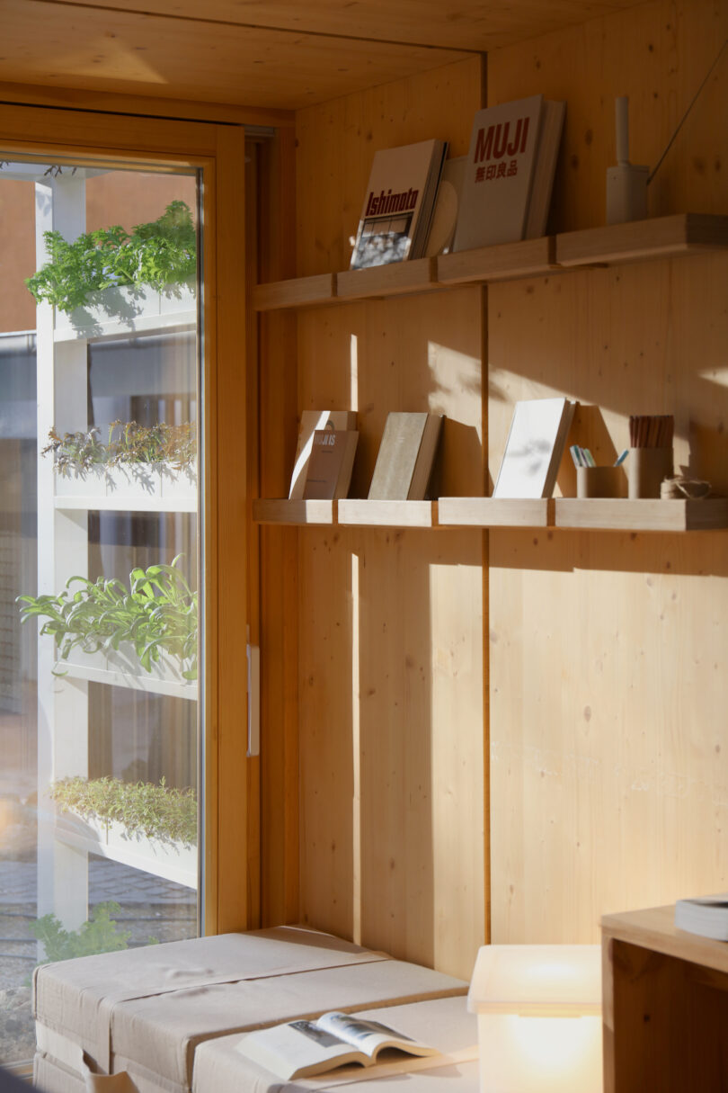 A small wooden reading nook with a bench, open book, shelves holding books and stationery, and a window showing greenery outside