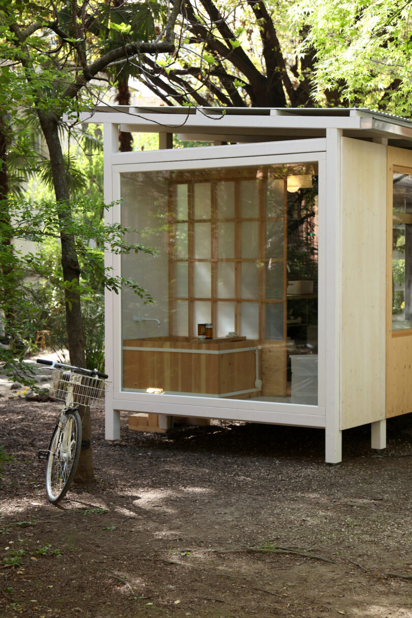 A small, modern wooden structure with large glass windows stands among trees; a bicycle with a basket is parked nearby