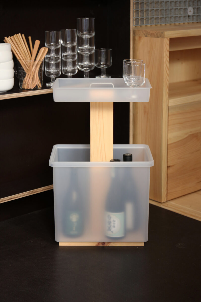 A two-tiered plastic storage bin with wooden support, holding bottles in the lower bin and a glass on the upper tray, placed next to glassware and bowls on shelves