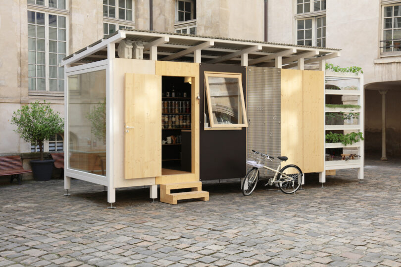 A small modern prefab structure with wood and glass panels sits on a stone courtyard, with a bike parked outside and vertical planters on one side
