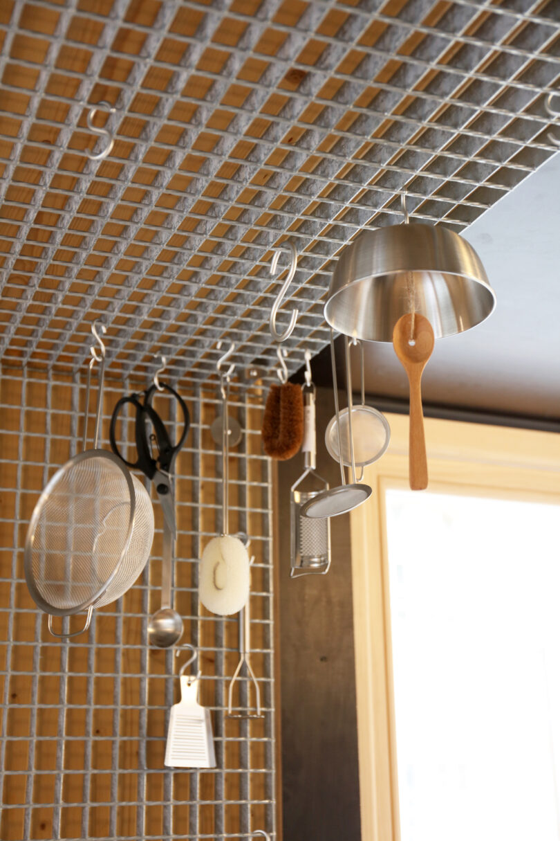 Kitchen utensils including a strainer, scissors, ladle, and sponge hang from hooks on a metal grid ceiling organizer near a window