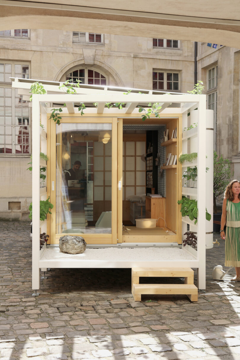 A small, modern, white tiny house with glass doors, plants growing on vertical trellises, and wooden steps, situated on a cobblestone courtyard. A woman stands nearby