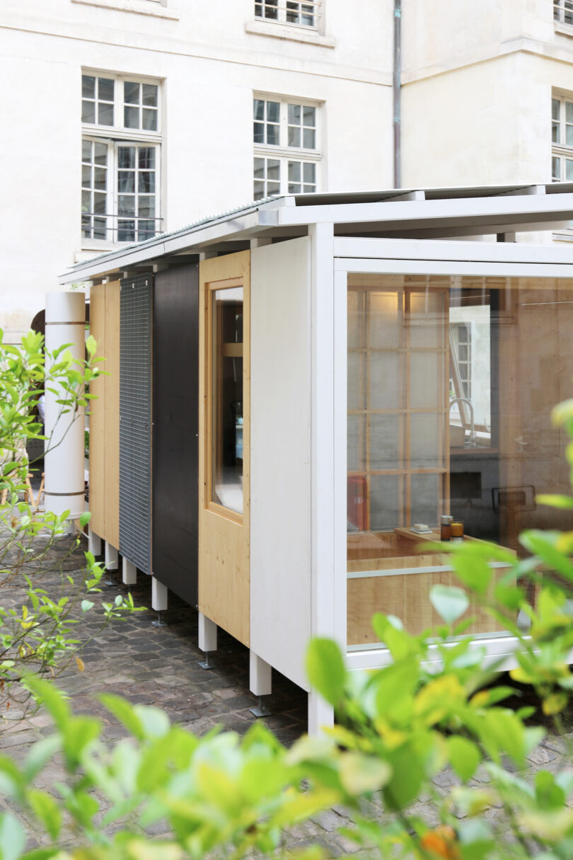 A modern, small modular building with large windows and a mix of wood and metal panels stands on an elevated foundation in a courtyard