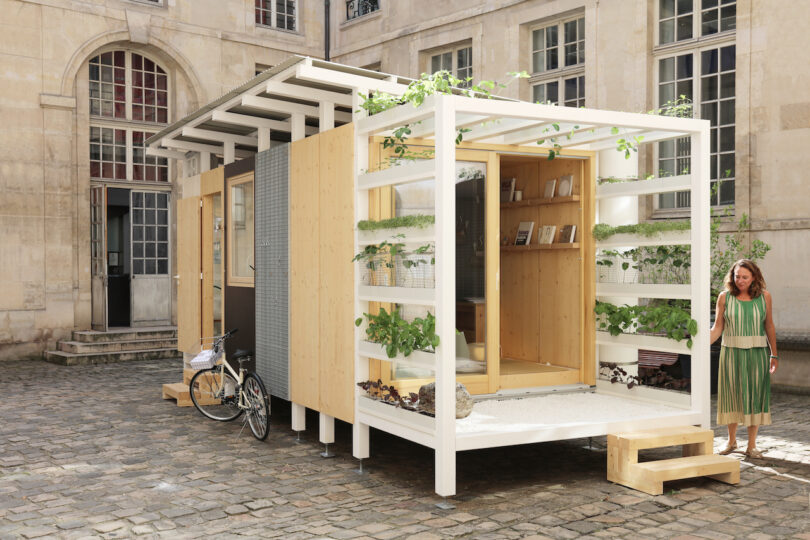 A small modern wooden tiny house with plants growing on the exterior, set in a stone courtyard; a woman stands on the front steps, and a bicycle is parked beside the house.