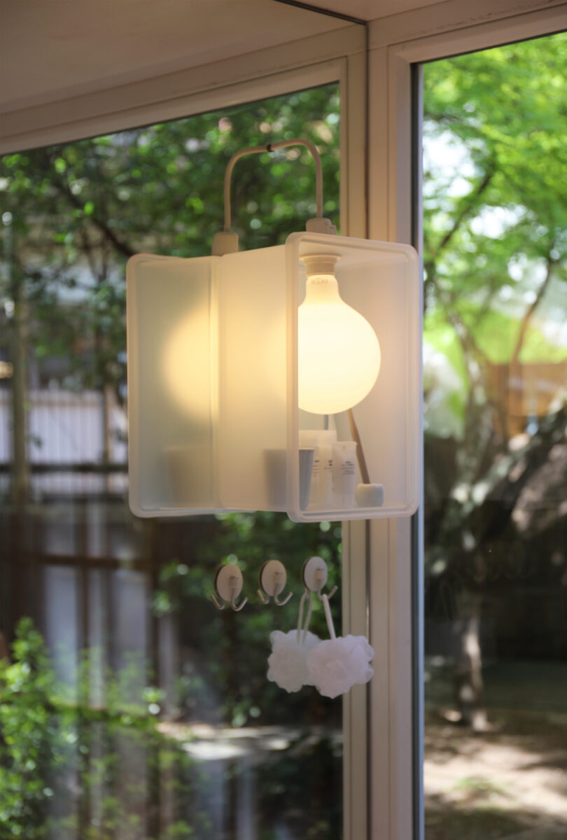 Frosted box-shaped light fixture with an exposed bulb hangs by a window, with hooks and a white bath pouf attached below. Green trees are visible outside