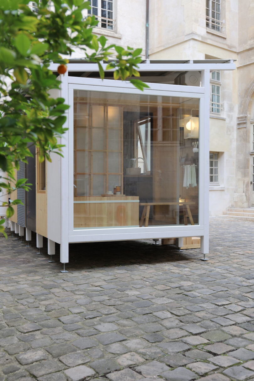 A modern, small glass-walled structure with a flat roof stands on a cobblestone courtyard, adjacent to a historic stone building
