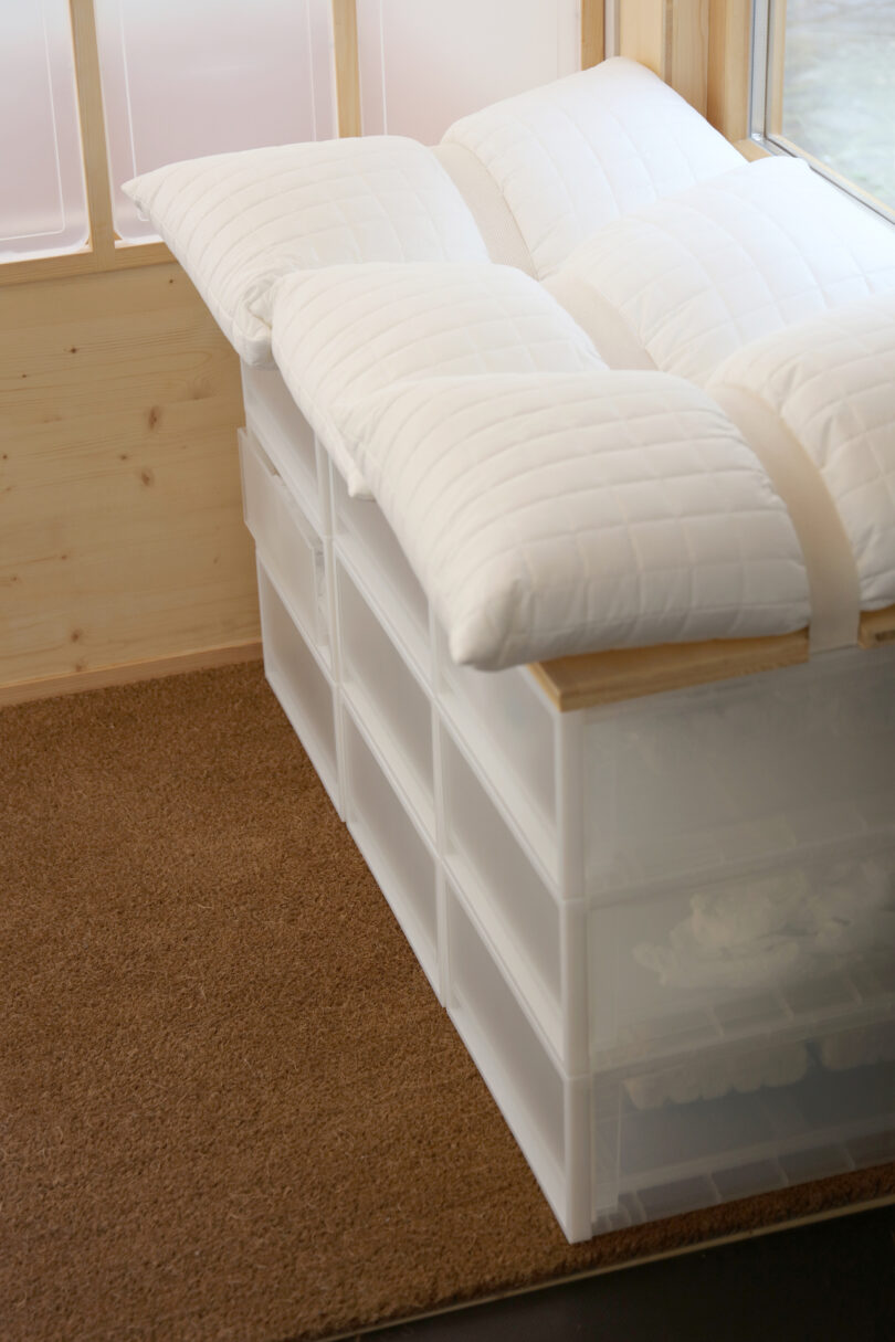 A low bench made of stacked white plastic storage drawers, topped with white pillows, is placed on a brown carpet next to a window