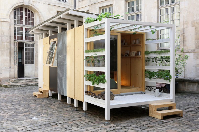 A modern tiny house with wood and metal exterior, glass doors, small porch, plants, and steps, situated in a stone courtyard beside an old building