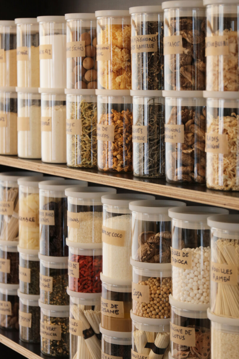 Plastic containers filled with various dried spices, herbs, and grains are neatly organized on shelves, each labeled with handwritten tags