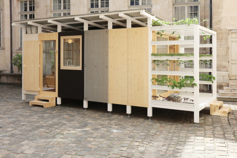A small modern wooden tiny house with a covered porch, large windows, and an attached vertical garden, situated on a cobblestone courtyard