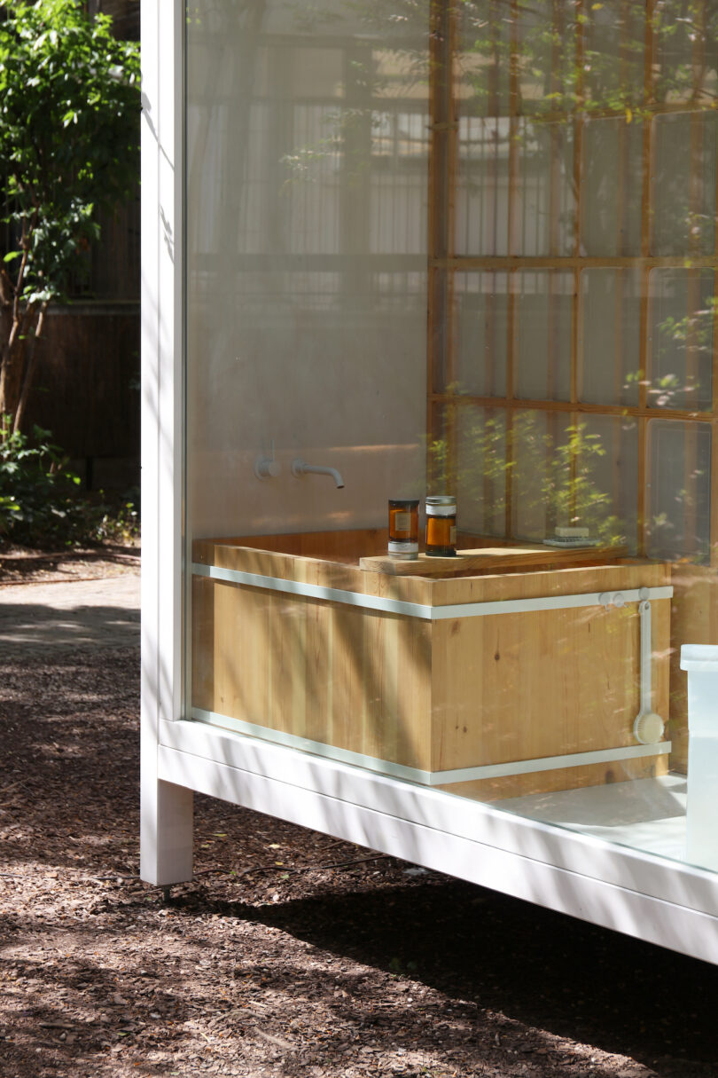 A wooden soaking tub and two bottles are inside a glass-walled structure with outdoor trees and sunlight visible through the windows