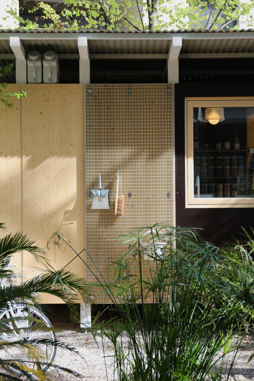 Outdoor wall with a wooden panel, grid section holding cleaning tools, a window with shelves visible inside, and plants in the foreground