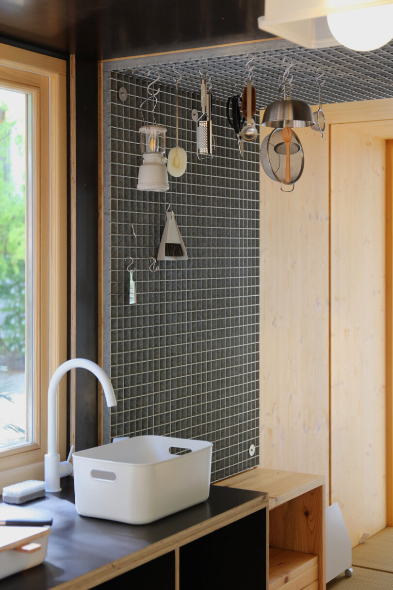 A modern kitchen corner with a white sink, faucet, black tiled backsplash, and various kitchen utensils hanging on a wall grid