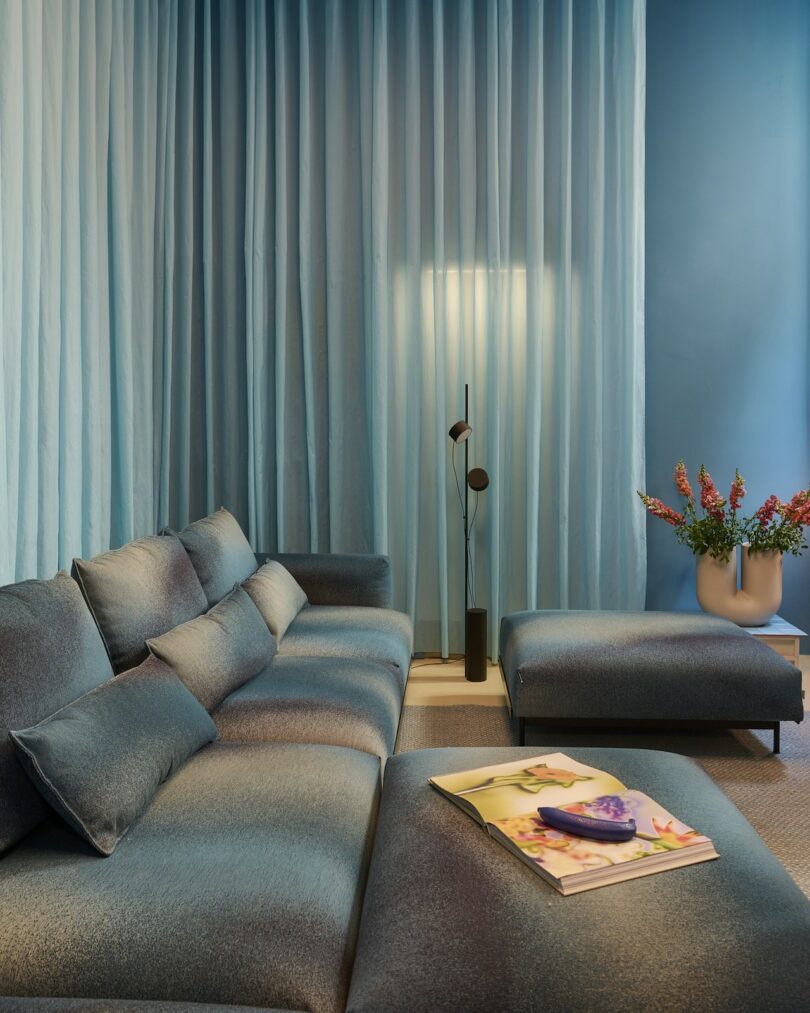 Modern living room with blue gradient sofa, matching ottoman, light blue floor-to-ceiling curtains, a black floor lamp, vase with flowers, and an open magazine on the ottoman