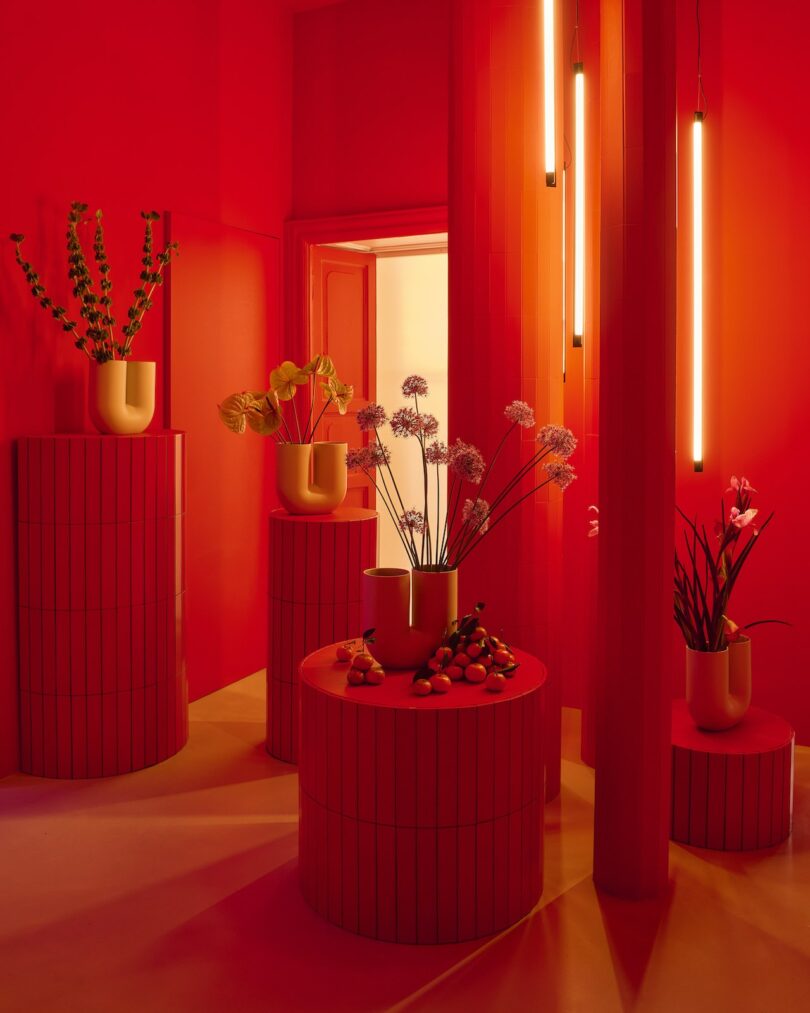A vibrant red room with cylindrical pedestals, yellow vases holding various flowers, hanging tube lights, and a display of assorted fruits