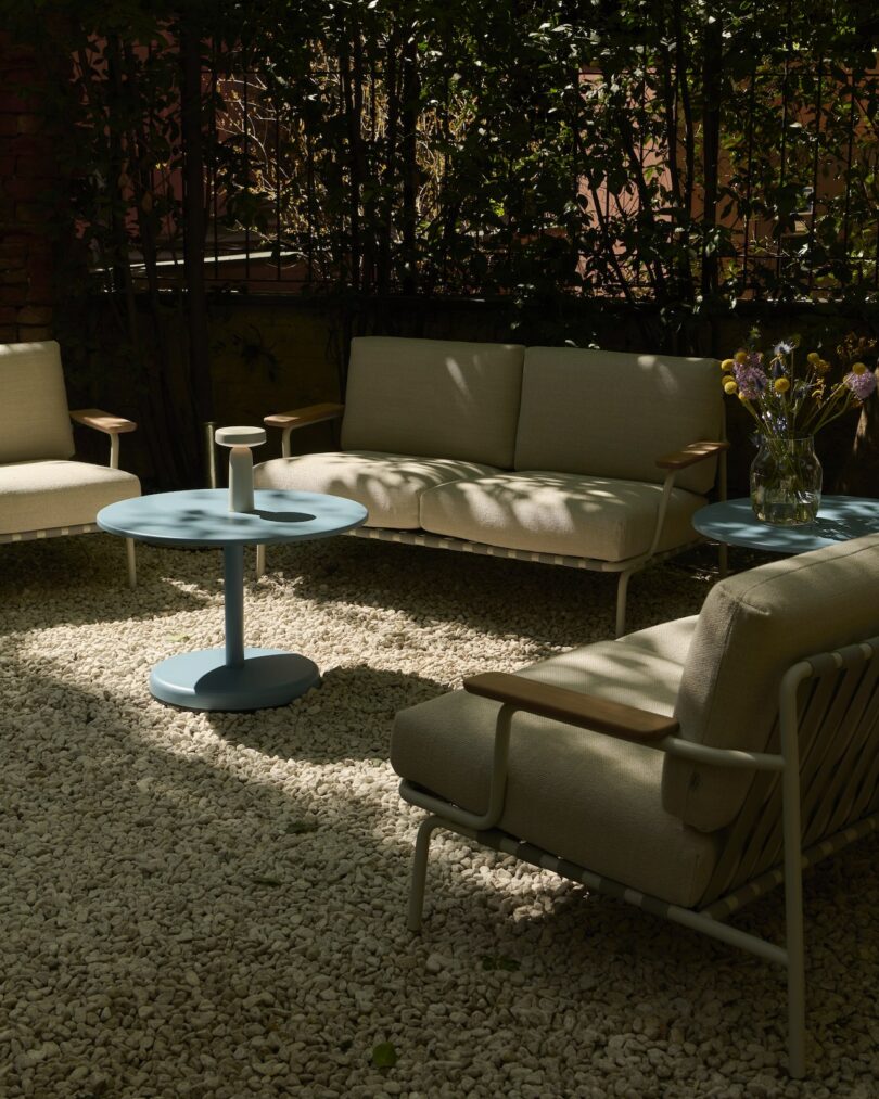 Outdoor seating area with beige cushioned chairs and sofas, two light blue tables, and a vase of flowers, set on white gravel with leafy plants in the background