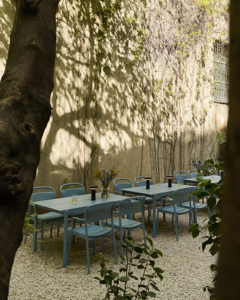 Outdoor patio with light blue tables and chairs set on white gravel, surrounded by greenery and a sunlit textured wall. Glasses and small vases are placed on the tables
